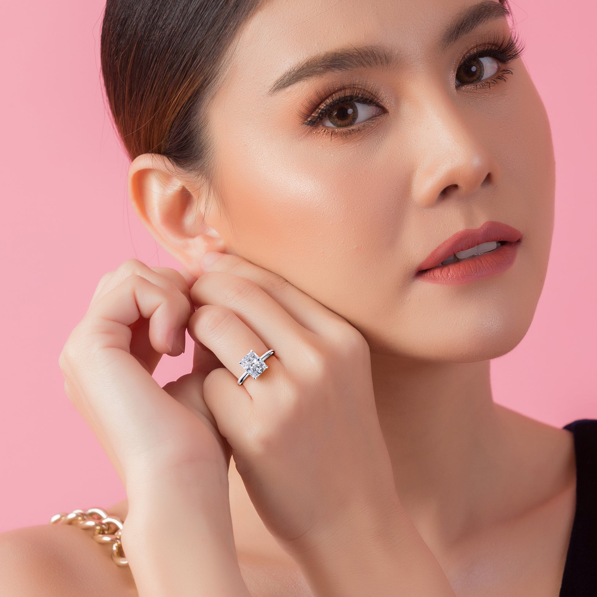 Woman wearing an elegant engagement ring with a radiant princess-cut diamond in platinum or 14K/18K white gold, on a soft pink backdrop.|
#14KRecycledWhiteGold
#18KRecycledWhiteGold
#Platinum