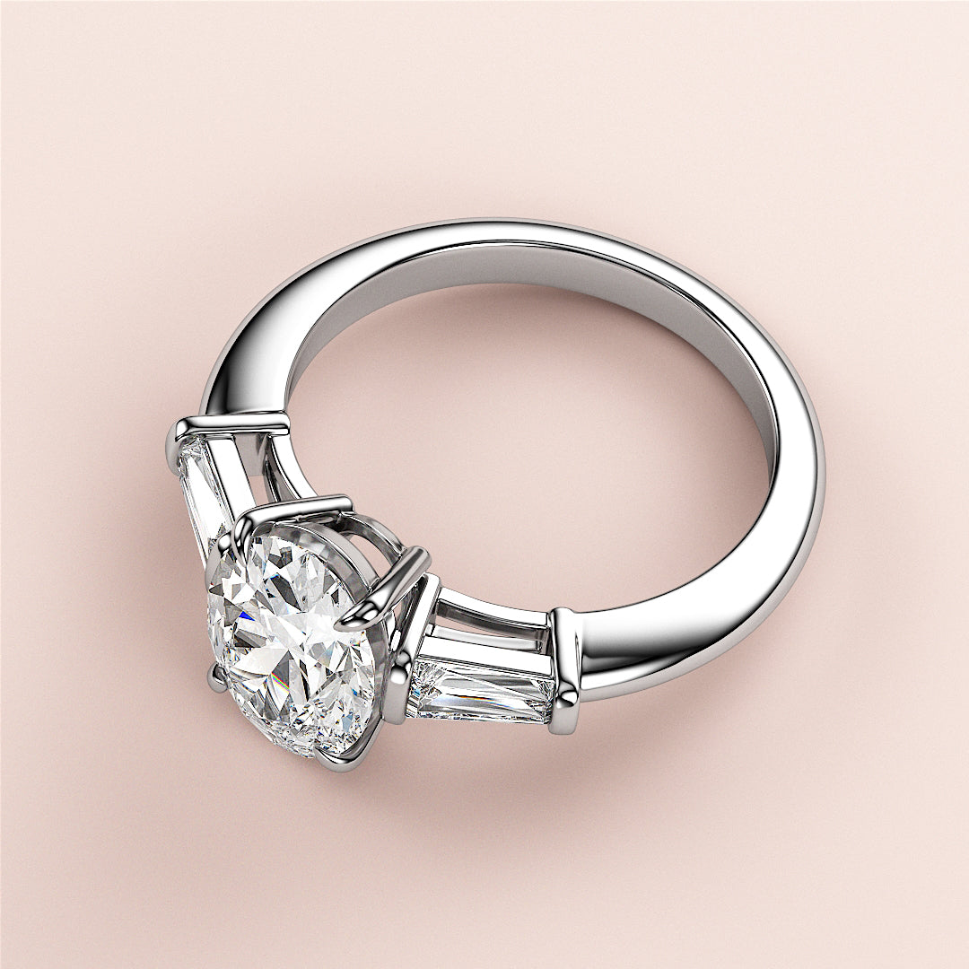 Elegant 14K white gold engagement ring with oval three-stone design and tapered baguettes, showcased against a soft pink background.|
#14KRecycledWhiteGold
#18KRecycledWhiteGold
#Platinum