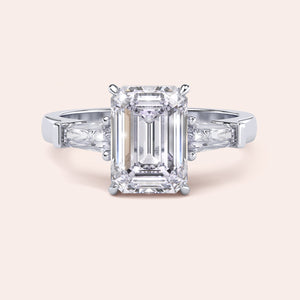 White gold engagement ring featuring a prominent emerald-cut diamond, flanked by smaller diamonds on a delicate band, exuding luxury. 