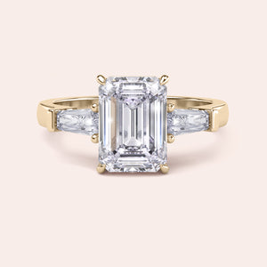 Yellow gold engagement ring featuring a prominent emerald-cut diamond, flanked by smaller diamonds on a delicate band, exuding luxury. 