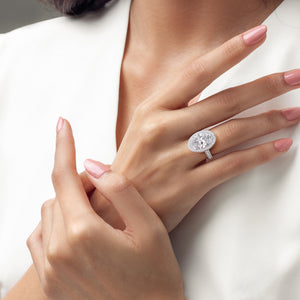 A woman's hand adorned with an oval diamond ring in 14K or 18K white gold, encircled by smaller diamonds, showcasing elegance.