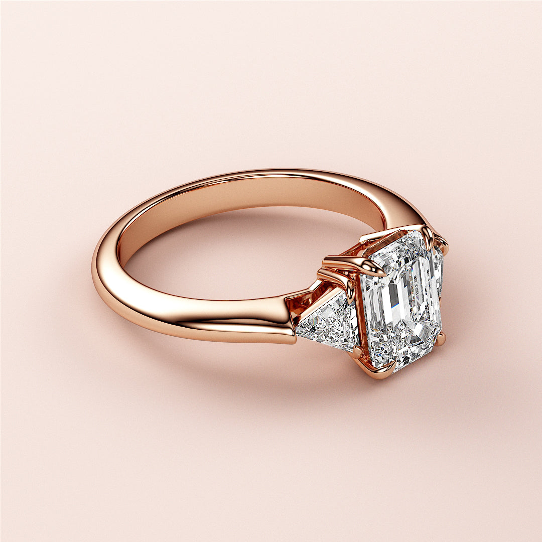 Elegant rose gold three-stone engagement ring in 14K or 18K gold featuring emerald-cut and triangular diamonds.