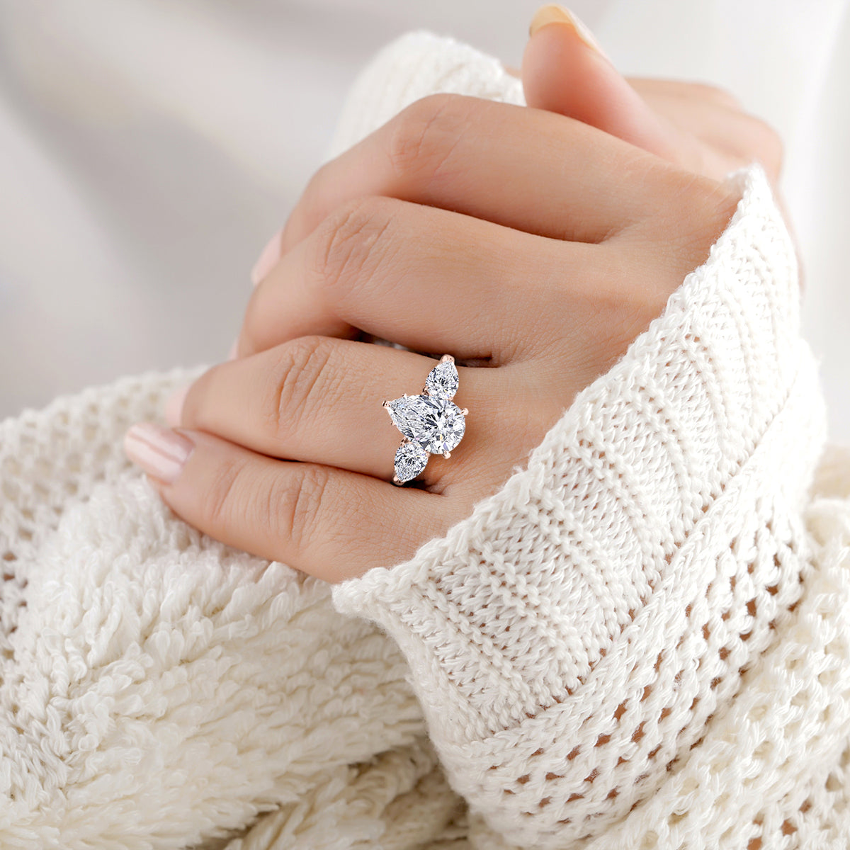 14K rose gold engagement ring featuring a pear-shaped diamond and side stones, worn on a woman's hand against white knit fabric.